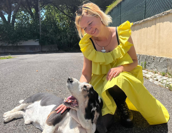 A woman in a yellow dress petting a basset dog on the street near a brick wall