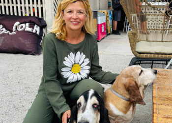 A woman in a green shirt petting a ginger basset dog named Rania and a black-and-white basset dog named Carol on the beach near a table